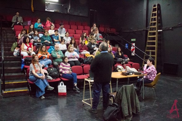 Imagen de un hombre de espalda, con un escritorio delante,  frente a un auditorio en gradas